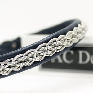 Puede incluir: Un brazalete de cuero azul marino con un diseño trenzado plateado. El brazalete está sobre una superficie blanca con una etiqueta en blanco y negro que dice "AC Design".