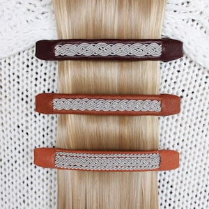 Op de afbeelding: Drie leren haarclips met zilveren gevlochten versiering. De clips zijn bruin, beige en donkerbruin.