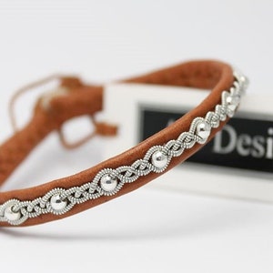 Könnte beinhalten: Ein braunes Lederarmband mit einem geflochtenen Silberdesign und silbernen Perlen. Das Armband liegt auf einer weißen Oberfläche mit einer weißen Karte, auf der in Schwarz "Design" steht.