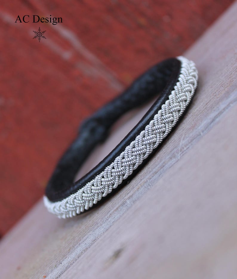 Sami bracelet Leipt handmade from Black reindeer leather and with a beautiful braid in pewter wire.
Shown against a red painted wall and a wooden floor.
AC Design and a little star in Black.