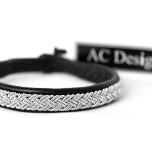 May include: A black leather bracelet with a silver braided design. The bracelet is tied with a knot and has a tag that reads "AC Design".