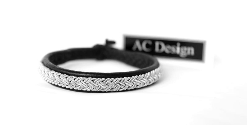 May include: A black leather bracelet with a silver braided design. The bracelet is tied with a knot and has a tag that reads "AC Design".