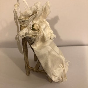 May include: A white fabric doll dressed in a white gown and lace sits on a small wooden chair. The chair is painted gold and has a simple design.