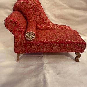 May include: Miniature red chaise longue with gold swirl pattern fabric. Features a rolled armrest with a decorative gold embellishment. The chaise has wooden legs and is designed for a dollhouse or miniature setting.