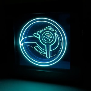 Puede incluir: Un letrero cuadrado iluminado con un diseño azul neón. El diseño presenta un emblema circular con un símbolo estilizado en el centro. El letrero brilla intensamente sobre un fondo oscuro, creando un efecto visual llamativo.