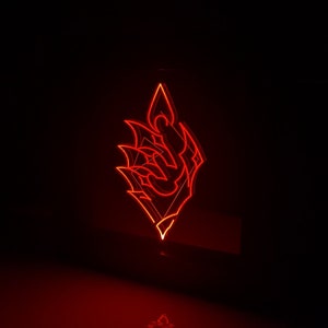 Puede incluir: Un letrero de neón rojo brillante en un entorno oscuro. El letrero tiene la forma de una llama o emblema estilizado, con ángulos agudos y una base puntiaguda. La luz se refleja en la superficie de abajo.