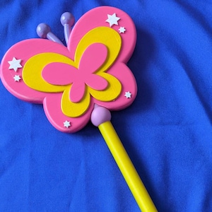 May include: A pink and yellow butterfly-shaped wand with a yellow handle and a pink ball at the end. The wand has white stars on it.