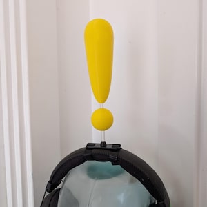 Puede incluir: Un soporte para auriculares en forma de signo de exclamación amarillo. El soporte está hecho de plástico y tiene una base redonda. El soporte está unido a unos auriculares negros.