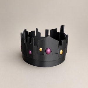 Può includere: Una corona nera caratterizzata da una silhouette frastagliata simile a un castello lungo il bordo superiore. La fascia è decorata con accenti circolari alternati in oro metallizzato e viola profondo scintillante, creando un netto contrasto con la superficie nera opaca.