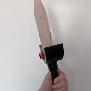 May include: A pink and black plastic sword with a red gem on the handle. The sword is held in a hand.