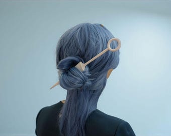 May include: A person's hair styled in a bun, secured with a light-colored hair stick. The hair is a shade of blue, and the person is wearing a black top. The hair stick has a geometric design.