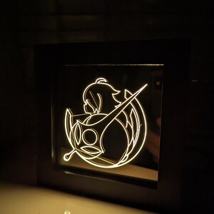 May include: A black framed light-up sign featuring a white silhouette of a woman holding a sword. The woman is wearing a long, flowing dress and has long hair. The sword is pointed upwards. The sign is illuminated from behind, creating a glowing effect.