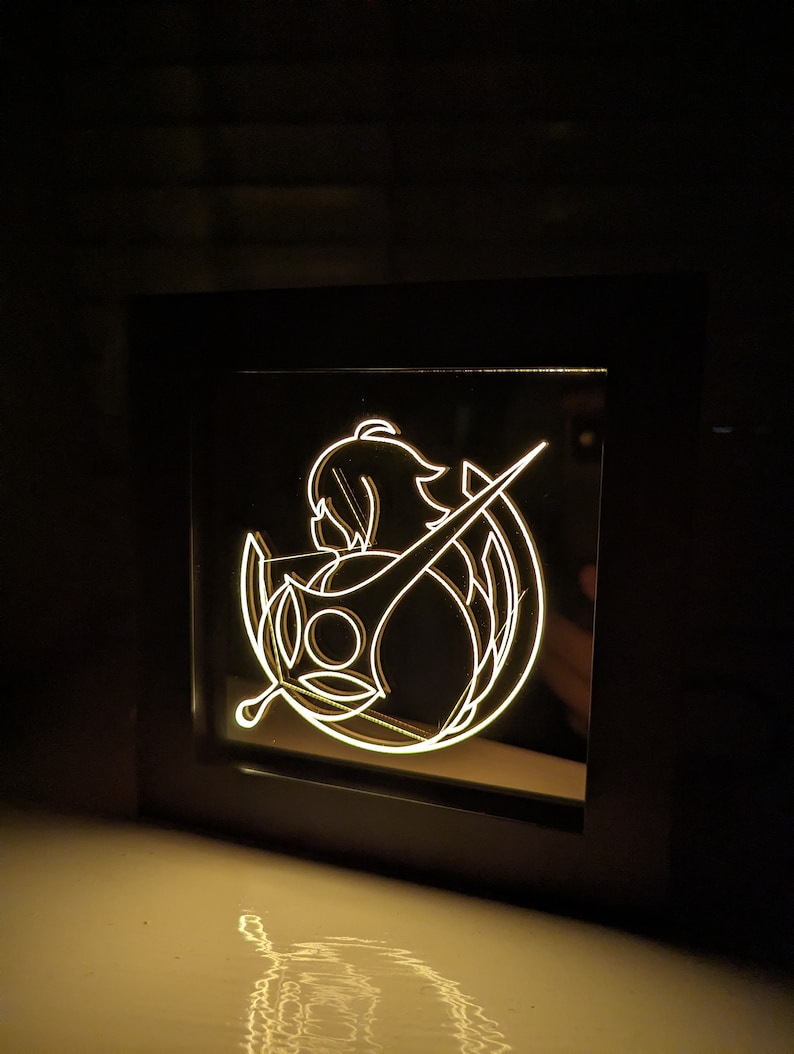 May include: A black framed light-up sign featuring a white silhouette of a woman holding a sword. The woman is wearing a long, flowing dress and has long hair. The sword is pointed upwards. The sign is illuminated from behind, creating a glowing effect.