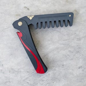 Peut inclure: Un peigne pliant noir et rouge avec un motif décoratif. Le peigne a un accent doré et un petit ouvre-bouteille.