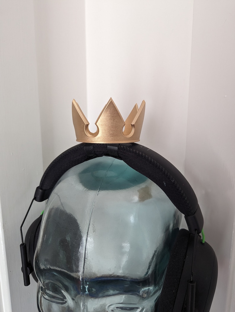 Pode incluir: Um suporte para fones de ouvido em forma de coroa de cor dourada. O suporte &eacute; feito de pl&aacute;stico e tem um acabamento liso e fosco. &Eacute; projetado para se ajustar &agrave; faixa de cabe&ccedil;a de um fone de ouvido.