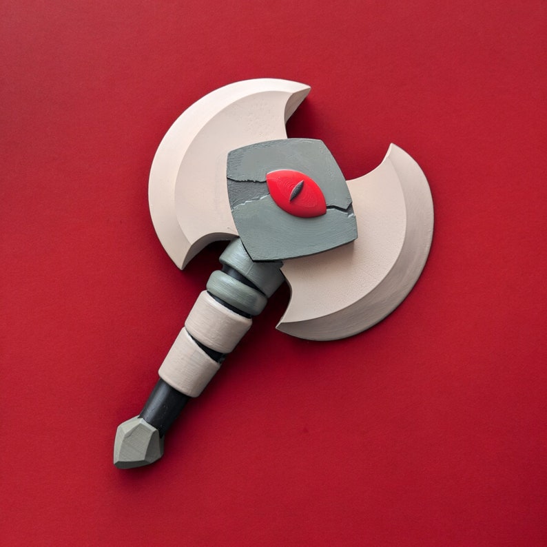 May include: A two-bladed axe with a stylized design. The axe head is light beige with a gray center featuring a red eye. The handle is black with beige and gray accents. The axe is set against a red background.