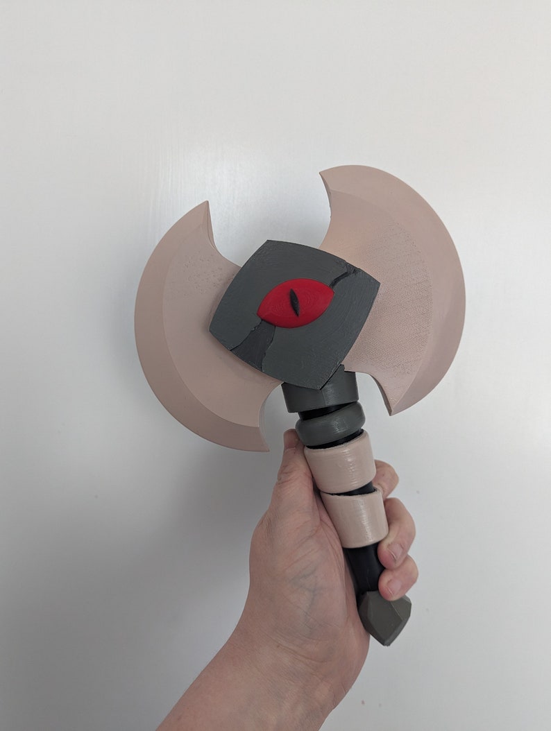May include: A two-handed axe prop with a light brown blade and a gray head featuring a red eye. The handle is black with tan and gray accents. The axe is designed for cosplay or display.