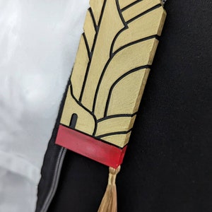 May include: A gold and red decorative pin with a stylized design and a tassel. The pin has a black outline and a red base.