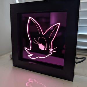 May include: A black framed LED light featuring a cartoon character's face with pink and white accents. The light is plugged in and sits on a white surface. The character's face is illuminated with a pink glow.