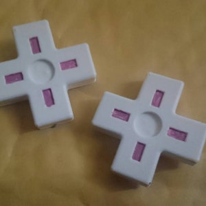 May include: Two white plastic cross-shaped objects with pink lines. The objects are on a brown surface.