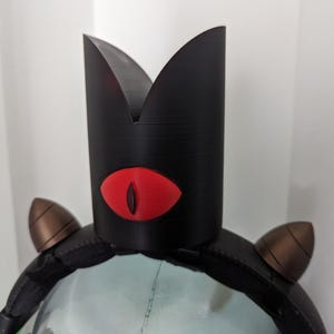 May include: A black crown-shaped headpiece with a red eye design. The headpiece has two brown, cone-shaped accents on the sides. The crown is made of a textured material and is worn on a black headband.