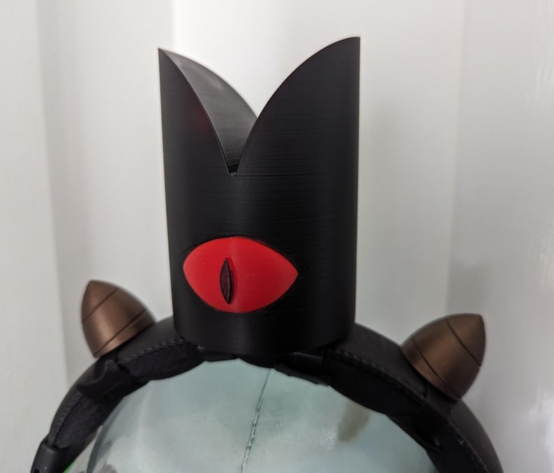May include: A black crown-shaped headpiece with a red eye design. The headpiece has two brown, cone-shaped accents on the sides. The crown is made of a textured material and is worn on a black headband.