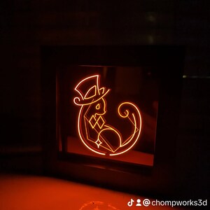 May include: A black framed light-up sign with an orange neon design of a cat wearing a top hat. The cat is sitting with its tail curled around its body. The sign is on a red surface and the text "@chompworks3d" is visible below the sign.