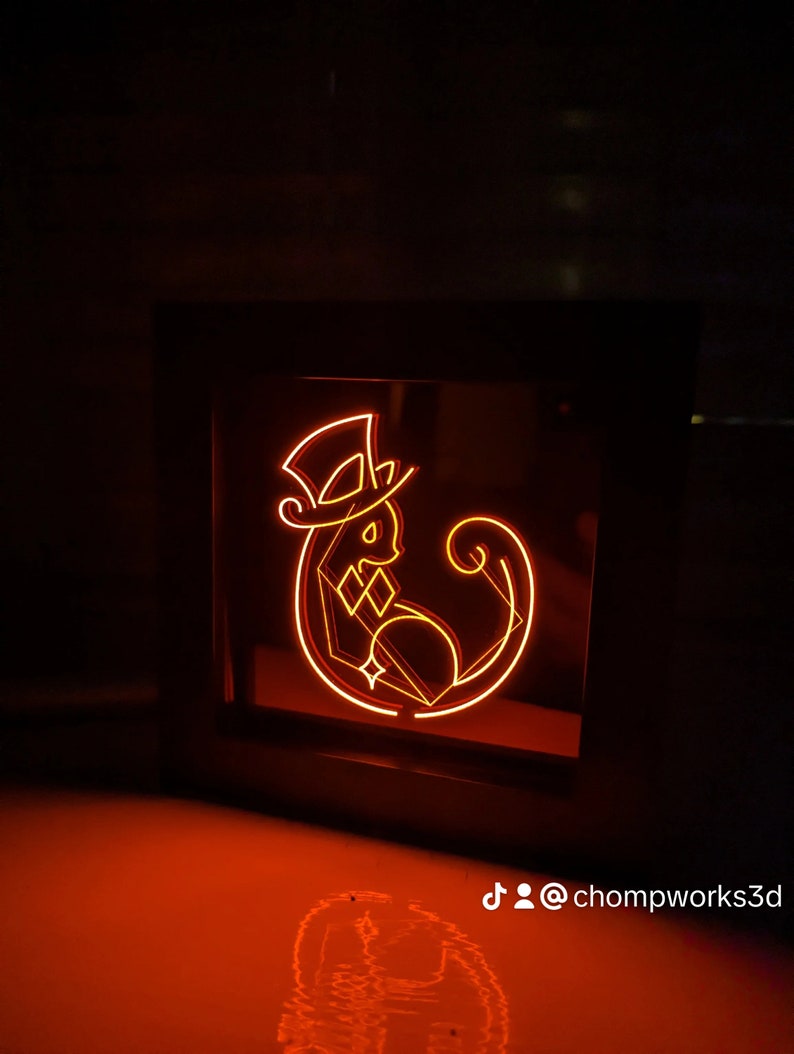 May include: A black framed light-up sign with an orange neon design of a cat wearing a top hat. The cat is sitting with its tail curled around its body. The sign is on a red surface and the text "@chompworks3d" is visible below the sign.
