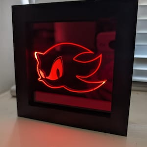 Puede incluir: Una obra de arte enmarcada e iluminada en negro, con el contorno rojo de la cabeza de un personaje. La obra está iluminada con un resplandor rojo, creando un efecto visual llamativo. El marco es cuadrado y se apoya sobre una superficie blanca.