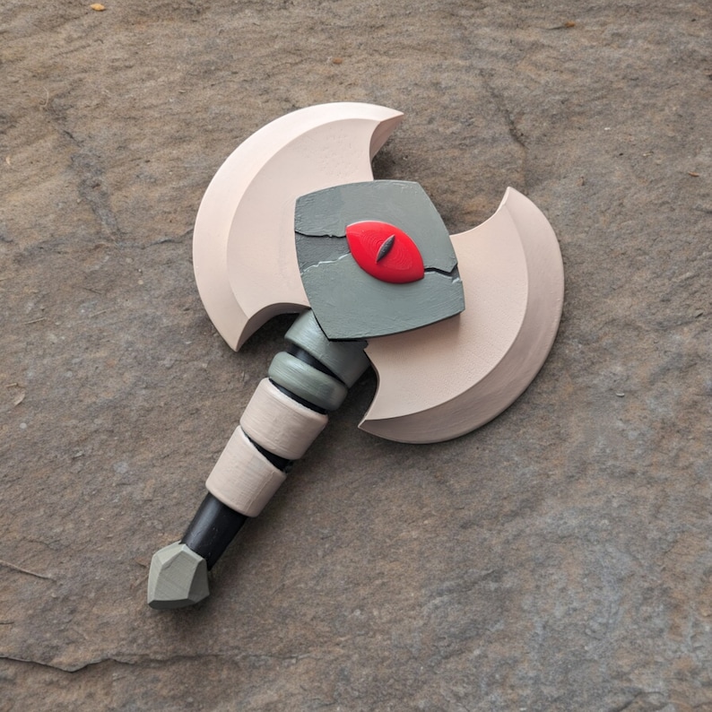 May include: A decorative axe with a light tan blade and a black handle. The axe head features a gray square with a red eye design. The handle is accented with light tan and gray rings.