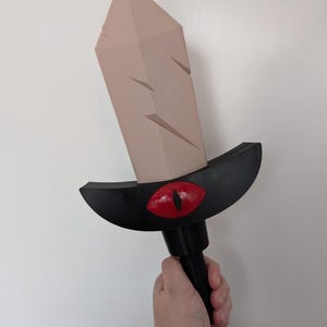 May include: A toy sword with a light brown blade and a black hilt. The hilt features a red eye design. The sword is held against a white background. The blade has several angled cutouts.