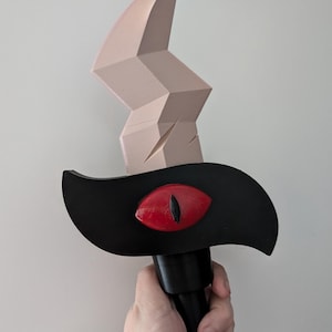 May include: A black and tan fantasy sword with a red eye design. The sword features a black handle, a black guard with a red eye, and a tan blade with a jagged design. The sword is held in a hand.