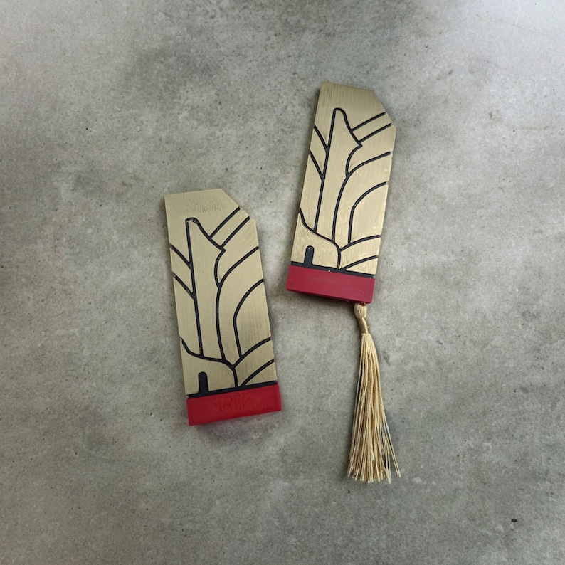 May include: Two gold bookmarks with red trim and black designs. The bookmarks feature a stylized image of a building with a tassel.