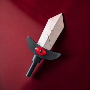 May include: A white sword with a black handle and a red eye design. The sword is made of paper and has a textured surface.