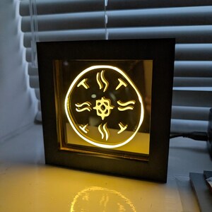 May include: A square, black-framed illuminated art piece. The artwork features a circular design with a central shape and surrounding curved lines, all glowing with a warm yellow light. The piece is sitting on a white surface.