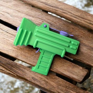 May include: A green plastic toy gun with a purple handle and a black trigger. The gun is lying on a wooden surface.