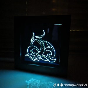 May include: A black framed light-up wall art piece featuring a blue and green neon design of a dragon. The design is intricate and detailed, with the dragon's body curled up in a protective pose. The light shines through the design, creating a beautiful and ethereal glow.
