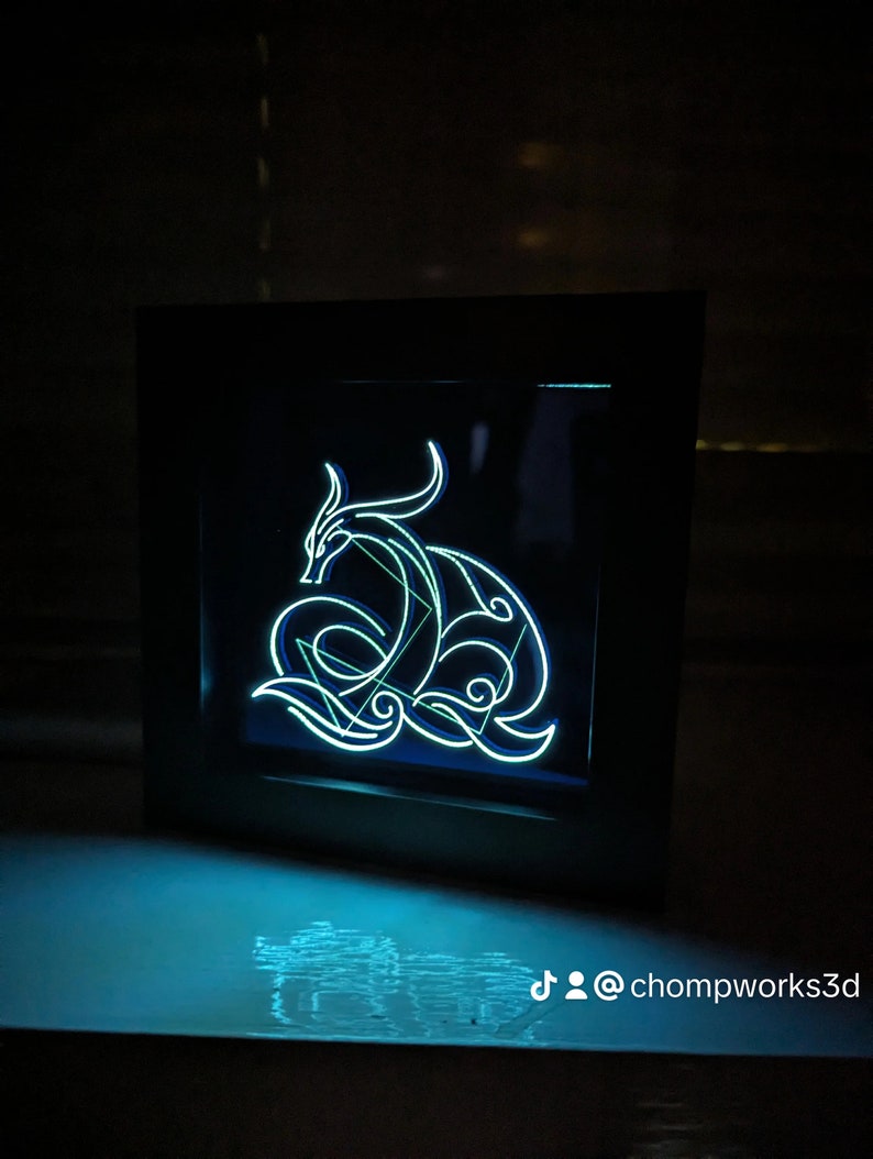 May include: A black framed light-up wall art piece featuring a blue and green neon design of a dragon. The design is intricate and detailed, with the dragon's body curled up in a protective pose. The light shines through the design, creating a beautiful and ethereal glow.