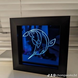 May include: A black framed 3D light sculpture of a stylised dolphin with a blue background. The light sculpture is lit up with a blue light. The TikTok handle @chompworks3d is visible in the bottom right corner of the image.