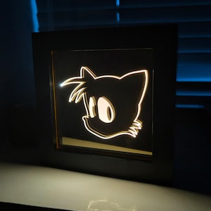 May include: A framed, illuminated artwork featuring the silhouette of a cartoon character's head. The character's face is outlined in a warm, glowing light against a black background. The frame is black, and the artwork is displayed on a white surface.