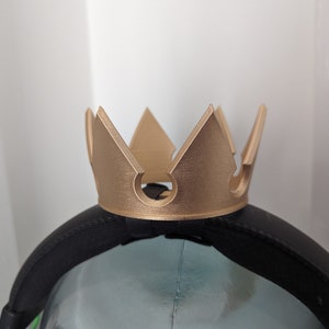Pode incluir: Uma faixa de cabe&ccedil;a de coroa dourada com um design pontudo. A faixa de cabe&ccedil;a &eacute; feita de pl&aacute;stico e tem uma faixa el&aacute;stica preta.