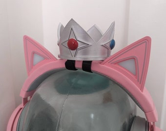 May include: A pink headset with cat ears and a silver crown. The headset has pink cat ears with white accents and lights. The silver crown has red and blue accents. The headset is on a clear, rounded surface.