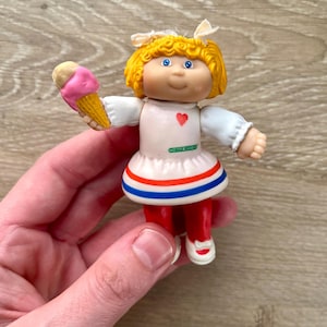 May include: A vintage Cabbage Patch Kids doll holding an ice cream cone. The doll has yellow hair, blue eyes, and is wearing a white dress with a red heart and blue and red trim. The doll is wearing red pants and white shoes.