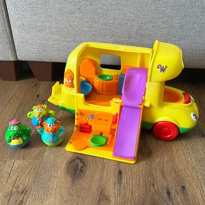 Può includere: Un camper giocattolo in plastica gialla con uno scivolo viola, caratterizzato da un interno aperto con mobili arancioni e un lavandino. Sono incluse tre piccole figure di animali colorate: un coccodrillo verde e due orsi con abiti blu e verdi.