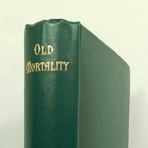 Puede incluir: Un libro de tapa dura verde con letras doradas. El título es "Old Mortality" de Sir Walter Scott. El libro está publicado por Black.