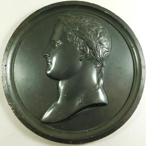 May include: A round, dark gray, metal medallion with a raised profile of a man's head in profile. The man is wearing a laurel wreath on his head and has short hair. The medallion has a raised rim around the edge.