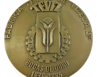 May include: A gold-colored medal with the text "Fabrica Textil de Vizela, Lda" and "Bodas de Ouro 1935-1985" around the edge. The center features a stylized image of a plant with a gear.