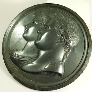 May include: A dark, circular relief sculpture featuring two profiles facing left. The figures are depicted in classical attire, with one wearing a laurel wreath. The piece has a weathered appearance, suggesting age and historical significance.