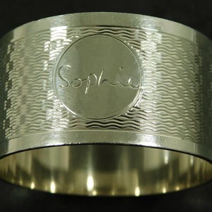 Könnte beinhalten: Ein silberfarbener Serviettenring aus Metall mit polierter Oberfläche und einem strukturierten Muster aus horizontalen Wellenlinien. In der Mitte des Rings befindet sich ein glattes, kreisförmiges Medaillon mit der Gravur Sophia in Schreibschrift.