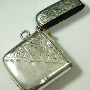 May include: A silver match safe with an engraved floral design. The match safe has a hinged lid and a small loop for attaching to a chain.
